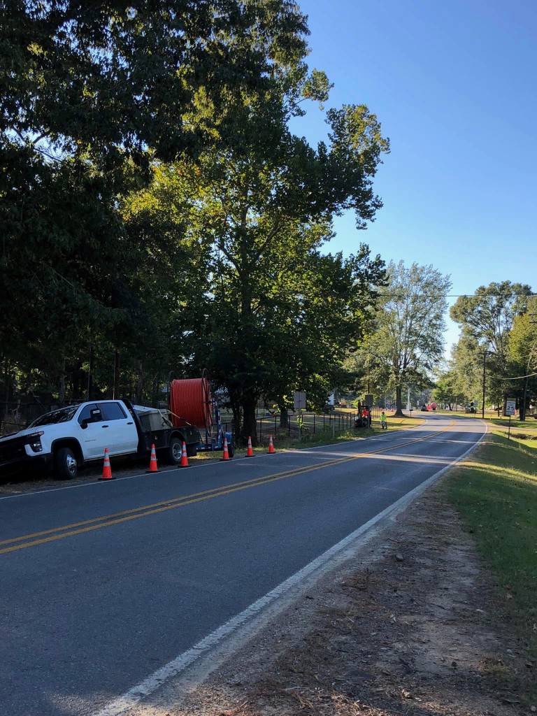 Utility truck towing orange conduit spool along a tree-lined road with safety cones